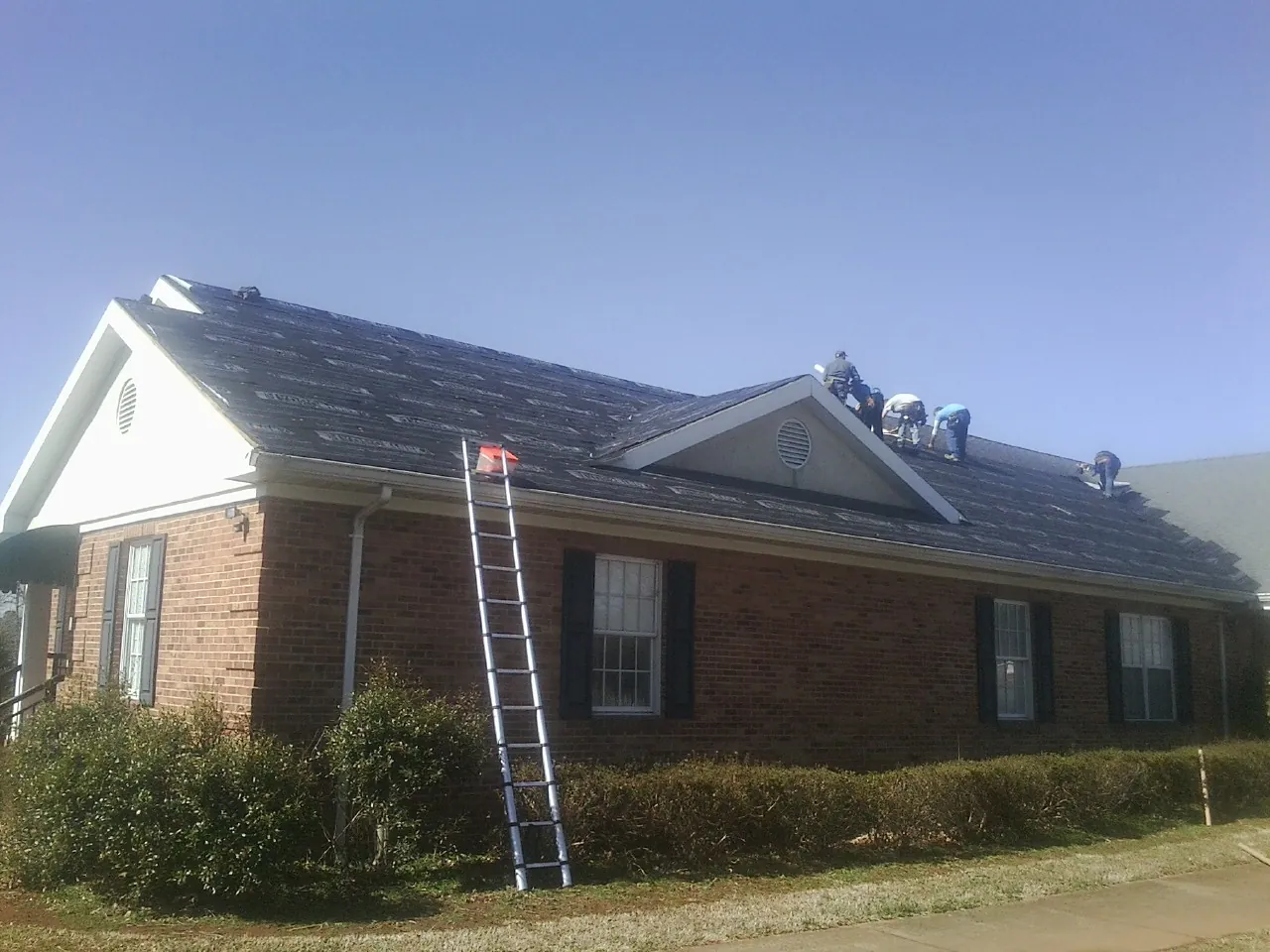 Crew tearing shingles off a brick ranch roof with a ladder set — Metal Roof Repair work by our crew in Bertram