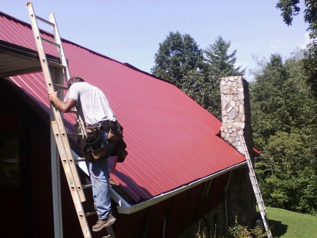 Roofer on a ladder beside a red metal roof and stone chimney — Soffit & Fascia Repair by Roofing Brothers in Bertram